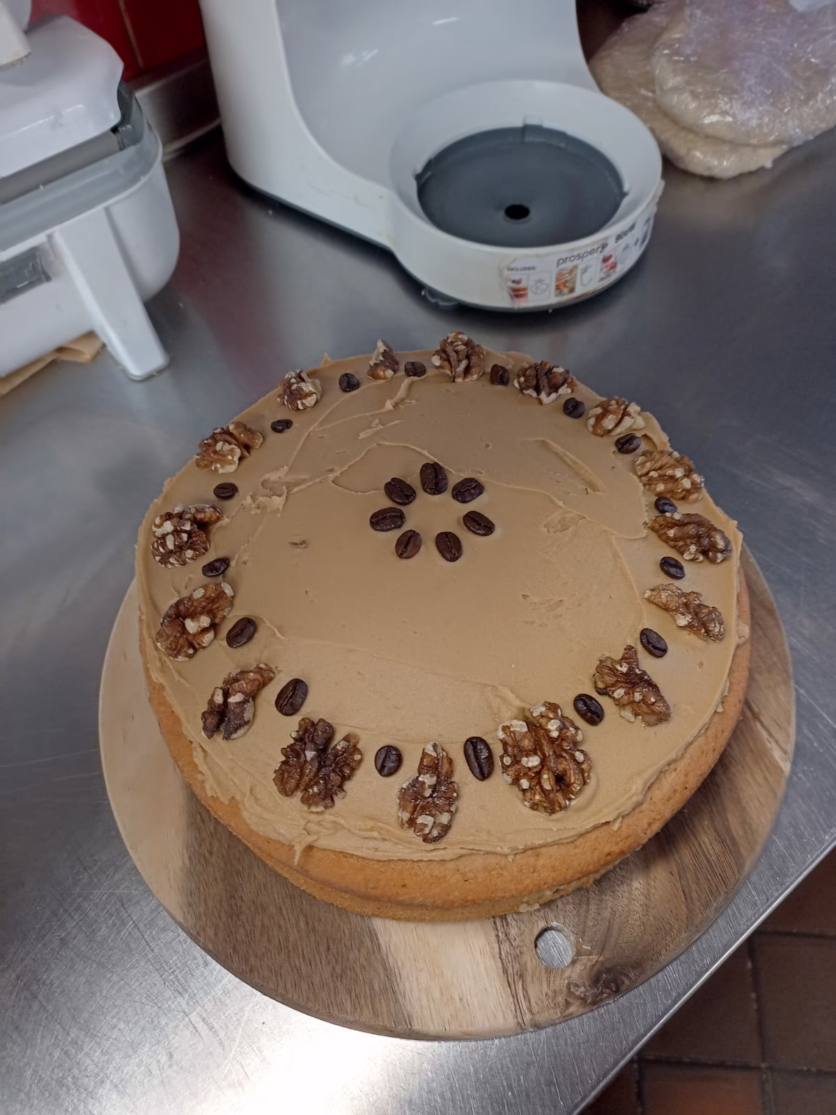 A homemade coffee and walnut cake topped with smooth coffee icing, decorated with whole coffee beans and walnuts, sitting on a wooden board in a kitchen.