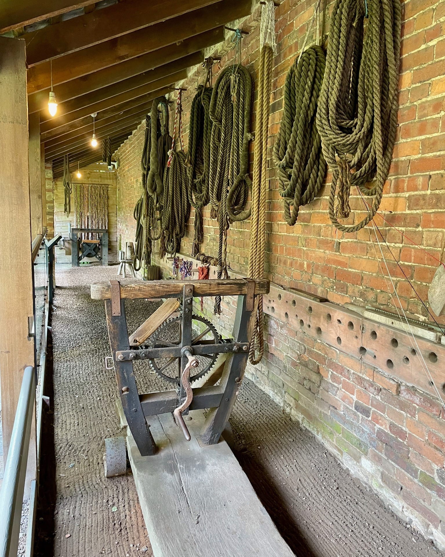 A perspective view down a covered outdoor corridor lined with red brick. In the foreground stands a vintage wooden rope-twisting machine with iron gears. Various thicknesses of heavy hemp rope are coiled and hanging from hooks along the brick wall on the right.
