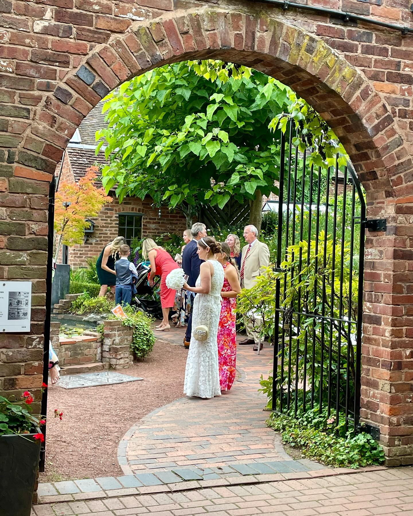A view through a historic brick archway and wrought iron gate into the Herb Garden at Bewdley Museum, where wedding guests are gathered. A bride in a white lace dress holds a bouquet, surrounded by friends and family in vibrant attire. The garden is lush with greenery, with a tree providing shade and a small pond visible in the background. The setting blends natural beauty with rustic charm, creating a picturesque wedding scene.