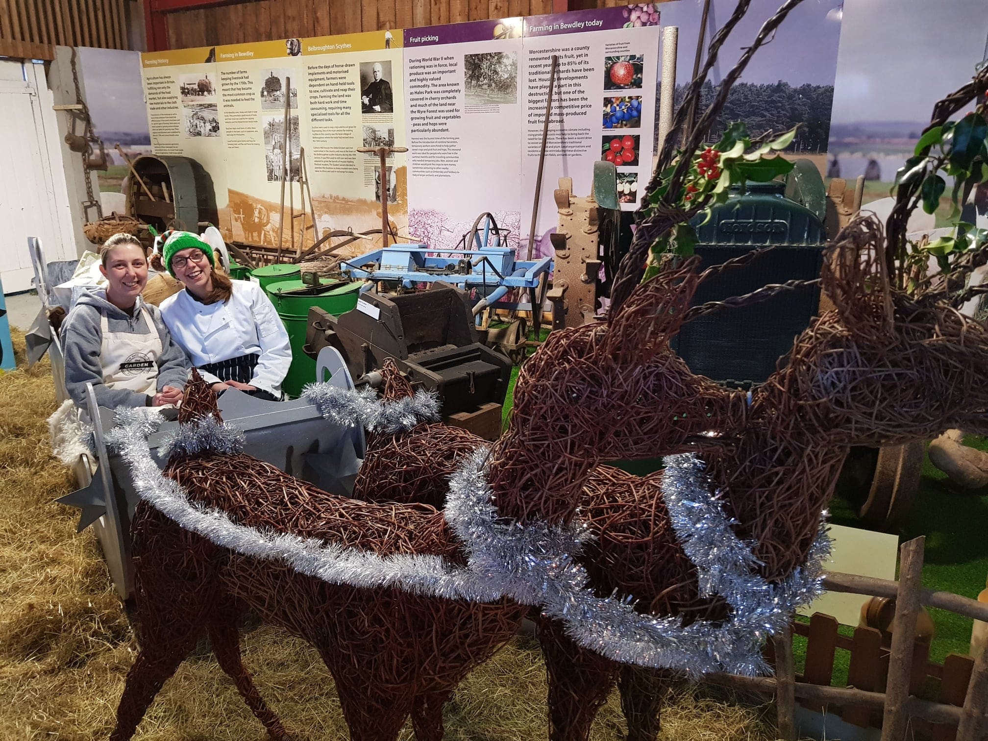 Garden Kitchen team member Lulu and owner Sarah smiling broadly while sitting in a Christmas sleigh led by wicker reindeer. They are posed in the covered Saw Yard area of the Museum.