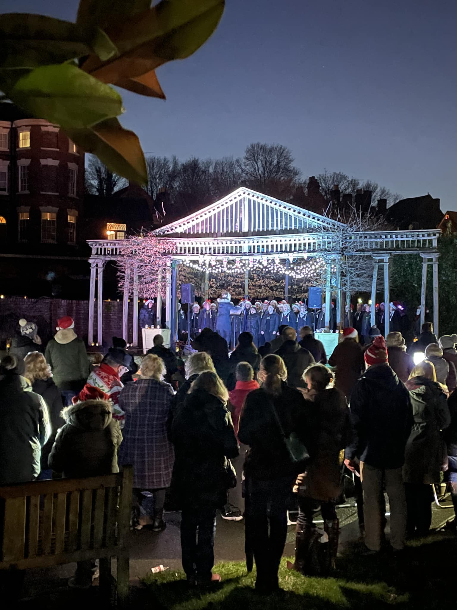 Carols by Candlelight in Jubilee Gardens, Bewdley Evening crowd gathered in Jubilee Gardens, Bewdley, watching a choir perform on a festively lit bandstand during the Carols by Candlelight event.
