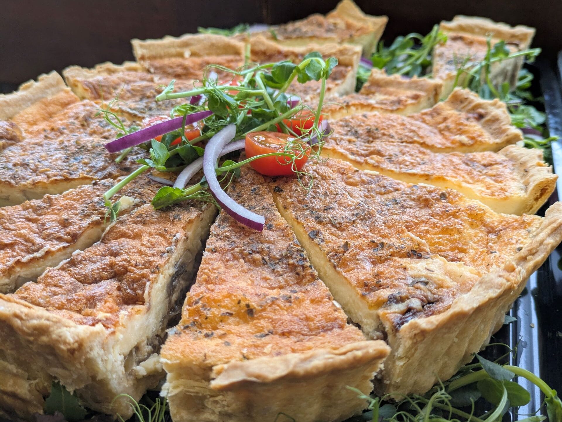 A close-up image of a sliced quiche displayed on a tray, garnished with fresh salad leaves, pea shoots, red onion, and cherry tomato halves, with natural light coming through a wooden window in the background.