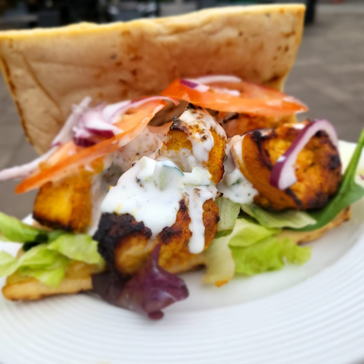 Chicken Tikka Flatbread with Fresh Salad and Yoghurt Dressing Close-up of a Chicken Tikka Flatbread, filled with marinated grilled chicken, fresh lettuce, tomato, red onion, and topped with cucumber and mint yoghurt dressing. Served on a white plate with an outdoor café setting in the background.