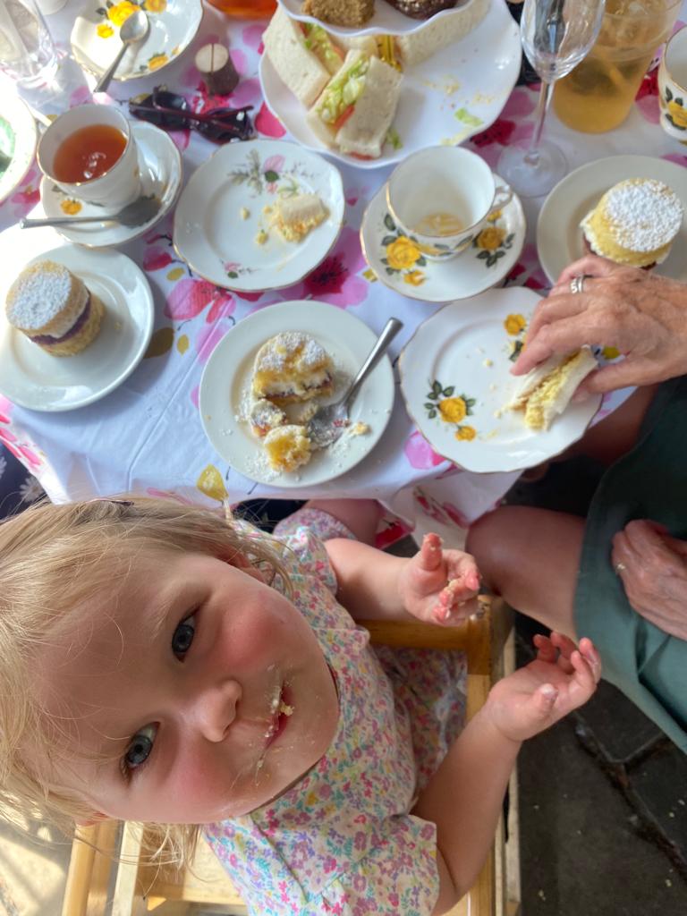 Young child with cake on her face smiling at the camera, seated at a table with sandwiches, scones, cakes, and vintage china during afternoon tea.
